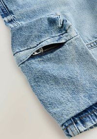 Veste en denim bleu clair avec une poche zippée sur la manche, au design à poignets et avec des détails de couture visibles.
