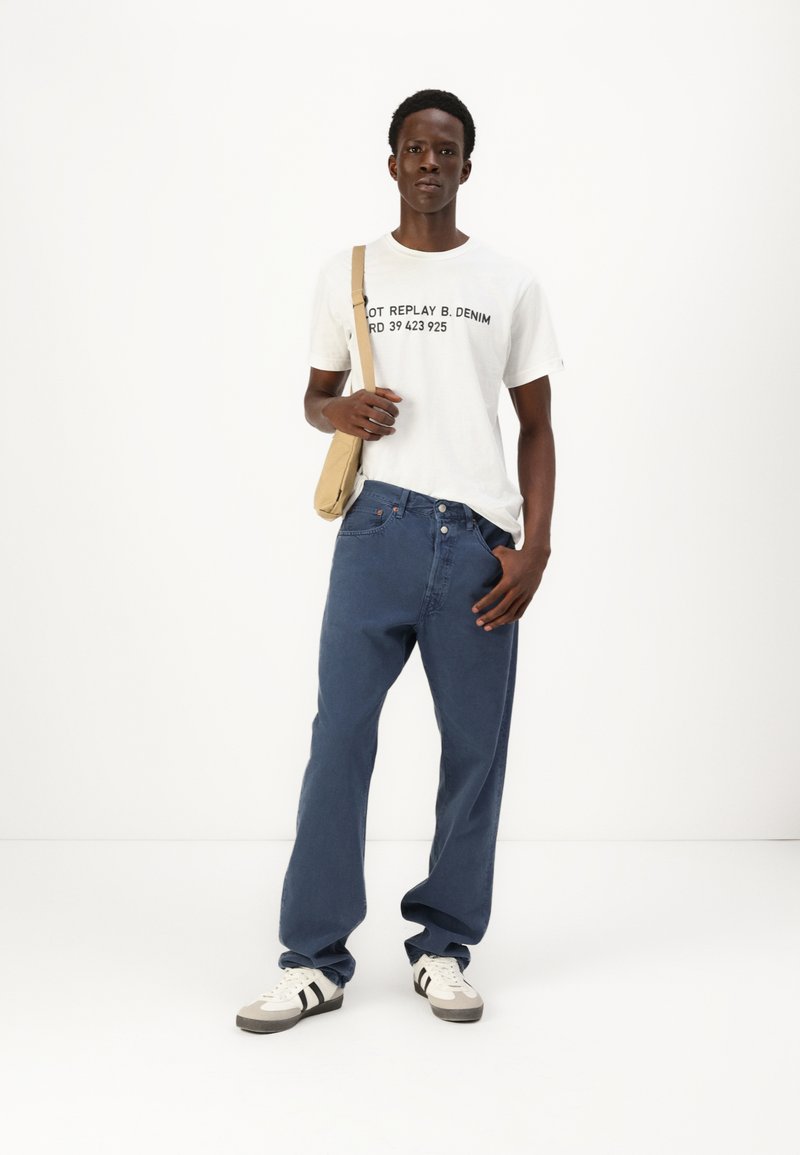 T-shirt blanche avec du texte noir, jean bleu ample, sac bandoulière beige et baskets blanches avec des rayures noires. Tenue simple et décontractée.