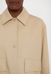 Beige ülekaaluline jakk, millel on struktureeritud krae, kahel esiküljel taskud ja nööbiga sulgemine, valmistatud siledast, tekstuurilisest kangast.