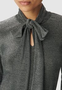 Blusa metallica nera e argento con dettaglio al collo a fiocco. Apertura a V e tessuto testurizzato con un leggero lucido.