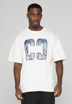 Man draagt een oversized wit T-shirt met grote blauw gemarmerde letters "C" en "J" op de voorkant, staand tegen een effen lichte achtergrond.