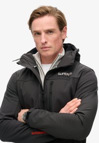 Zwart geruite jas met hoge kraag, ritssluiting aan de voorkant en logo-accenten. Beschikt over een grijze fleece voering en een getailleerd ontwerp.