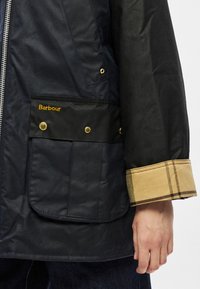 Tengerészkék viaszolt kabát, fekete zsebrátétellel, arany színű szerelékkel, barna kockás mandzsetta részlettel és oldalsó cipzáras záródással. A márka logója látható.