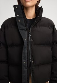Giacca a vento nera con design trapuntato, colletto alto, zip frontale e dettagli con bottoni a pressione, realizzata in tessuto liscio e strutturato.