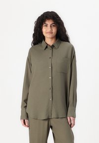 Camisa verde-oliva com botões, com colarinho pontudo, mangas longas e um único bolso no peito. Feita de um tecido leve e macio.