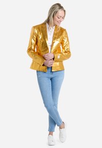 Blazer de oro metálico con un corte ajustado, que presenta un cierre de un solo botón y solapas en pico, combinado con jeans ajustados azul claro y zapatillas blancas.