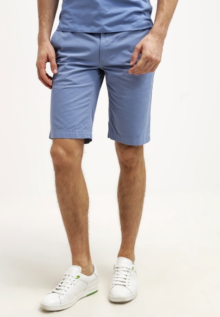 Homme portant un short bleu clair jusqu'aux genoux, une chemise bleue assortie et des baskets blanches, debout sur un fond clair uni.