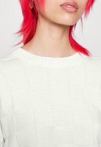 Vit stickad tröja med en texturerad rutmönster och en ribbad rundad halsringning. Slät och mjuk textur synlig i tyget.