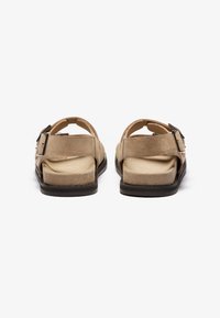 Sandales en daim beige avec des sangles réglables et une boucle en métal rectangulaire, dotées d'une semelle plate et d'un dos ouvert pour plus de confort.