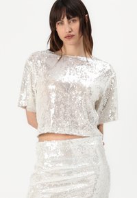 Top corto argento realizzato in tessuto di paillettes, con maniche corte e una texture a coste. Abbinato a una gonna in paillettes coordinata.