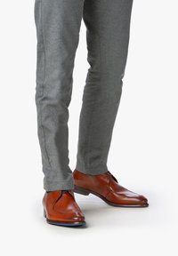 Bottines en cuir marron avec une finition lisse, associées à un pantalon en tissu gris ajusté présentant une texture subtile et un détail de couture latérale.