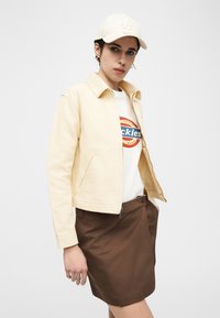 Άτομο που φοράει εκρού καπέλο του μπέιζμπολ, ανοιχτό μπεζ μπουφάν, λευκό μπλουζάκι με γραφικό σχέδιο και καφέ φούστα, στέκεται μπροστά σε απλό λευκό φόντο.