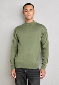 Pulover verde din tricot cu guler înalt, manșete și tiv cu nervuri. Textură moale, croială lejeră, asortat cu blugi negri. Design simplu.