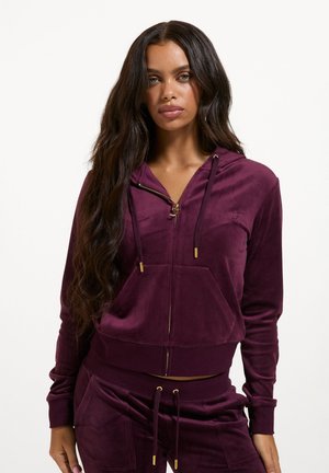 Frau mit langen dunklen Haaren, die eine lila Velour-Kapuzenjacke mit Reißverschluss und passende Kordelzughose vor schlichtem Hintergrund trägt.