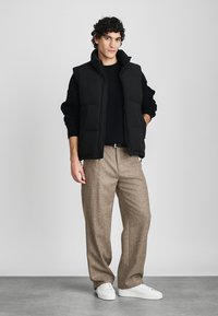 Svart pufferväst med hög krage över en mörk stickad tröja, kombinerad med ljusbruna strukturerade byxor och vita sneakers.