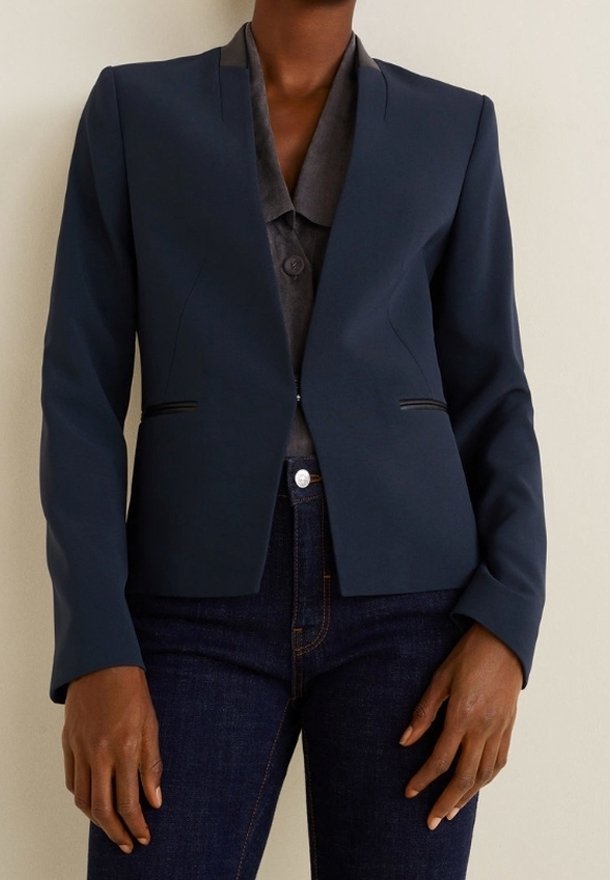 Personne portant un blazer bleu marine ajusté sur une chemise grise boutonnée et un jean bleu marine, debout devant un fond clair uni.