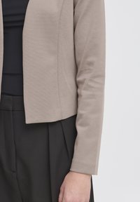 Veste beige à manches longues portée sur un haut noir et un pantalon noir plissé, bras et main détendus sur le côté.