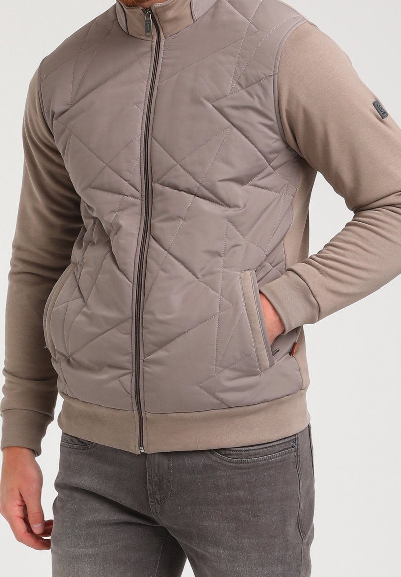 Homme portant une veste matelassée taupe à fermeture éclair avec manches longues et poches latérales, associée à un jean gris, debout avec une main dans la poche.