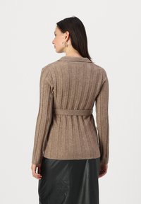 Brauner gerippter Cardigan mit einem Taillengürtel, langen Ärmeln und einem Kragen, getragen über einem schwarzen Rock. Weiche Textur, minimale Details.