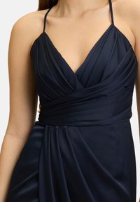 Granatowa sukienka wieczorowa z dekoltem w kształcie litery V i cienkimi ramiączkami, z drapowanym materiałem i dopasowaną talią. Gładka tekstura i elegancki fason.