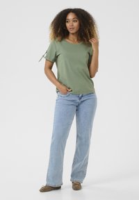 Olive green, short-sleeved t-shirt with a round neckline and side tie detail, paired with light blue straight-leg jeans and brown flats.