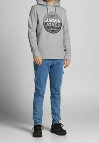 Felpa grigia con cappuccio e stampa grafica nera, abbinata a jeans blu e sneakers nere. Design semplice, materiale in cotone e vestibilità rilassata.