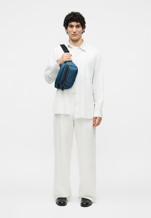 Mladý muž s kučeravými vlasmi nosí voľnú bielu košeľu a nohavice, drží modrú vzorovanú crossbody tašku s čiernym popruhom a zipsom.