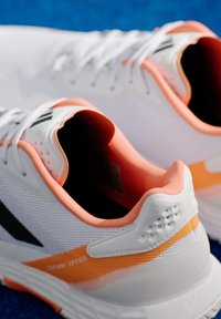 Λευκά αθλητικά παπούτσια με πορτοκαλί λεπτομέρειες, υφή πλέγματος στην επάνω πλευρά, μαξιλαρωμένος γιαλός και μαύρο λογότυπο. Διαθέτουν ζωντανή αντικραδασμική προστασία και κομψό σχέδιο.