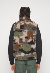 Chaleco de forro polar marrón, verde y beige con un patrón de camuflaje, que presenta un cuello alto y una superficie suave y texturizada.