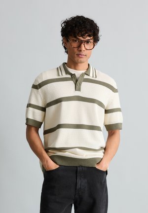 Junger Mann mit lockigem Haar und Brille, der ein weißes, gestricktes Poloshirt mit olivgrünen Streifen und schwarze Hosen trägt, die Hände in den Taschen.