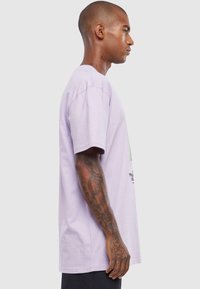 Lavender bomulds T-shirt med korte ærmer, lige skæring, med grøn grafisk tekst på fronten og tatoveringer synlige på armen.