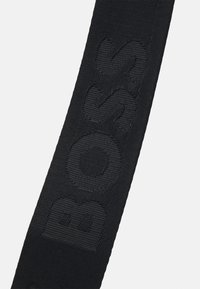 Czarny materiałowy pasek z tłoczoną teksturą logo "BOSS", charakteryzujący się gładką, tkany teksturą, odpowiedni do torebek lub akcesoriów.