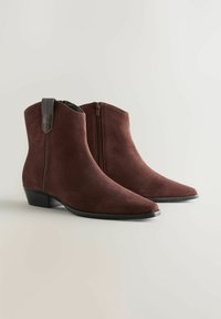Un par de botas de tobillo de ante marrón oscuro con cremallera lateral y tacones bajos en un fondo claro.
