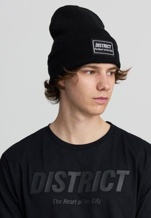Beanie in maglia nera con un patch logo che presenta il testo "DISTRICT" e "Il Cuore della Città." Indossato con una t-shirt nera, vista laterale in primo piano.