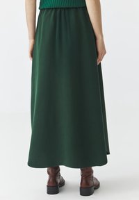 Lange groene rok met een gladde textuur en een geplooide tailleband, gecombineerd met bruine enkellaarsjes met een vierkante hak.