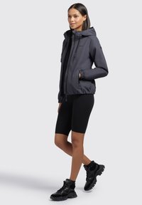 Veste zippée gris foncé avec capuche, poignets élastiques et deux poches avant, portée sur un haut noir et un short de cyclisme noir. Baskets noires.