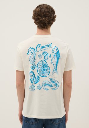 Mann trägt weißes T-Shirt mit blauen Skizzen von Meerestieren und Muscheln sowie dem Schriftzug „Cannes plage du Midi“ auf dem Rücken.