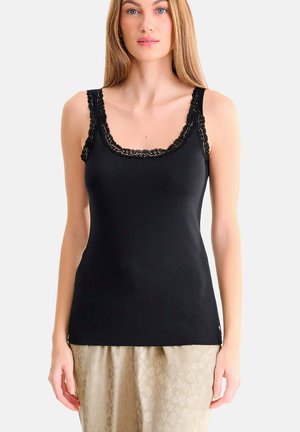 Femme portant un haut noir sans manches avec bordure en dentelle et un pantalon beige à motifs, debout devant un fond clair uni.
