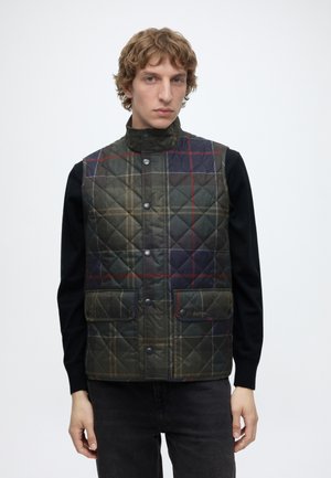 Junger Mann trägt eine gesteppte, geknöpfte, dunkelgrüne und marine karierte Weste über einem schwarzen Langarmshirt vor einem einfarbigen weißen Hintergrund.