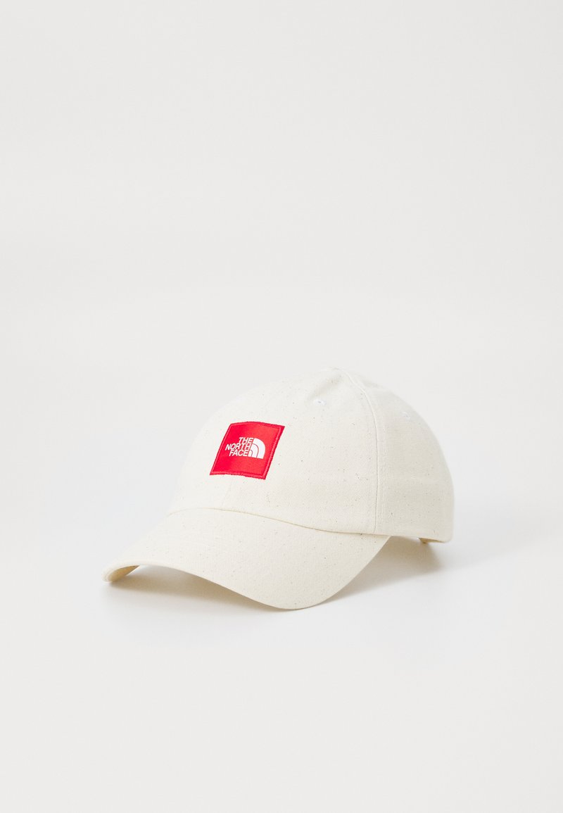 Gorra de béisbol color crema con visera curva, que presenta un parche rectangular rojo con un logo blanco. Fabricada en tela texturizada.