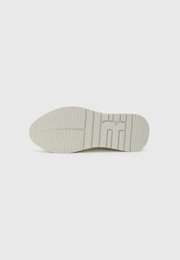 Sål af en sko, der har et let beige gummimateriale med vandrette riller for bedre skridsikkerhed og et subtilt logo prægede nær hælen.
