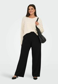Cremfarbener Kabelstrickpullover, lockere Passform; hochgeschnittene schwarze weite Hosen; schwarze Handtasche; spitze schwarze Schuhe mit Verzierungen.