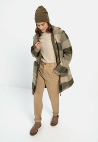 Karo-Mantel in Oliv und Beige, Strickmütze, beiges T-Shirt, tanfarbene Hose und braune Stiefeletten. Hat eine weiche Textur und eine lässige Passform.