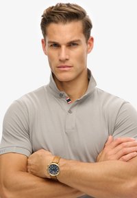 Lichtgrijze polo met een korte opstaande kraag, twee knopen en een gestreept detail bij de hals. Model draagt een bruine leren polshorloge.