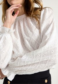 Hvid, tekstureret bluse med diamant- og blomster mønstre; lange ærmer med samlede manchetter; letvægtsstof. Båret med mørke bukser med knapper.