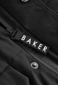 Juodas audinys su matinio paviršiaus apdaila, ant kurios yra balta etiketė su užrašu "BAKER", juodas sagtis ir metalinė spaudė. 