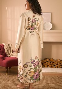 Raso floreale color crema, caratterizzato da fiori ricamati colorati e una cintura in vita. Maniche lunghe e design lungo fino alla caviglia con dettagli piping.