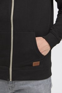 Crna zip-hoodica od mekanog materijala, s prednjim džepom, rebrastim rukavima i malom smeškom kožnom oznakom na donjoj strani.