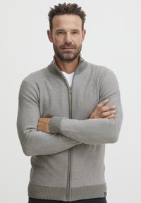 Szürke, zipzáros kardigán bordázott mandzsettákkal és aljjal, texturált kötött mintával. Magas gallérral és sima cipzárral rendelkezik.