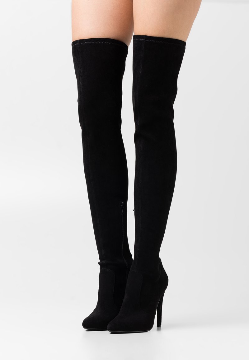 Botas de ante negras por encima de la rodilla con punta afilada, tacón alto de aguja y detalle de cierre lateral para facilitar su uso. Textura suave en toda la superficie.